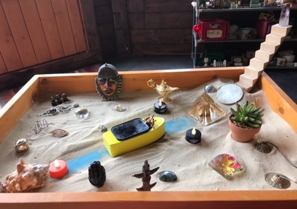 Sandtray featuring a boat figurine in the center of the tray pointed toward a set of stairs rising out of the tray. There are various figurines surrounding the boat.